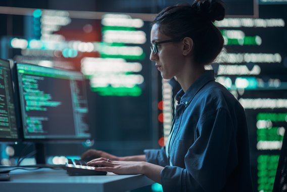 Vrouw werkt achter computer met programmeercode op meerdere schermen.