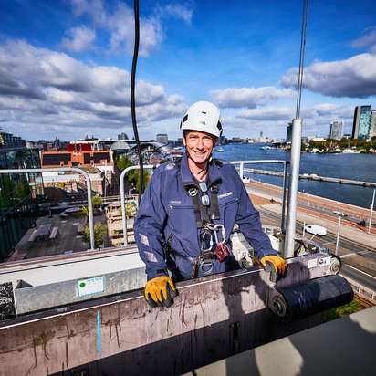 Vraag hier een offerte voor keuring gevelonderhoudsinstallatie aan TÜV NORD Inspecteur in een gevelonderhoudinstallatie met Amsterdam in de achtergrond.