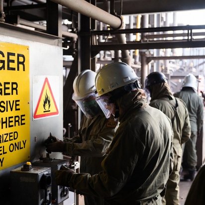 Medewerkers met beschermingsmiddelen werken bij industriële installatie met explosiegevaar-waarschuwingsbord.